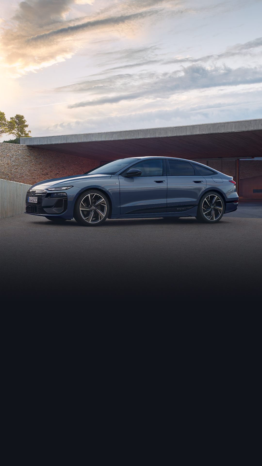 The A6 Sportback e-tron is parked in front of a house's carport, seen from the side. A young woman is standing next to it.