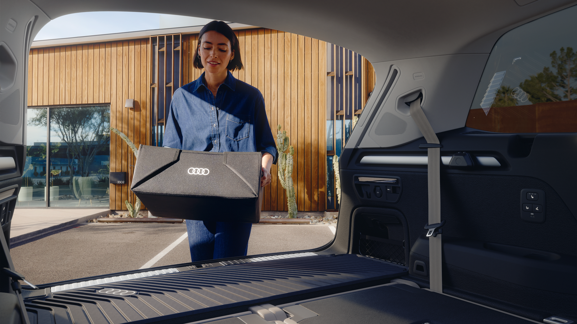 Vue depuis la banquette arrière sur le coffre ouvert, avec un gros plan sur le tapis de coffre. Une jeune femme y pose une petite boîte arborant le logo Audi.