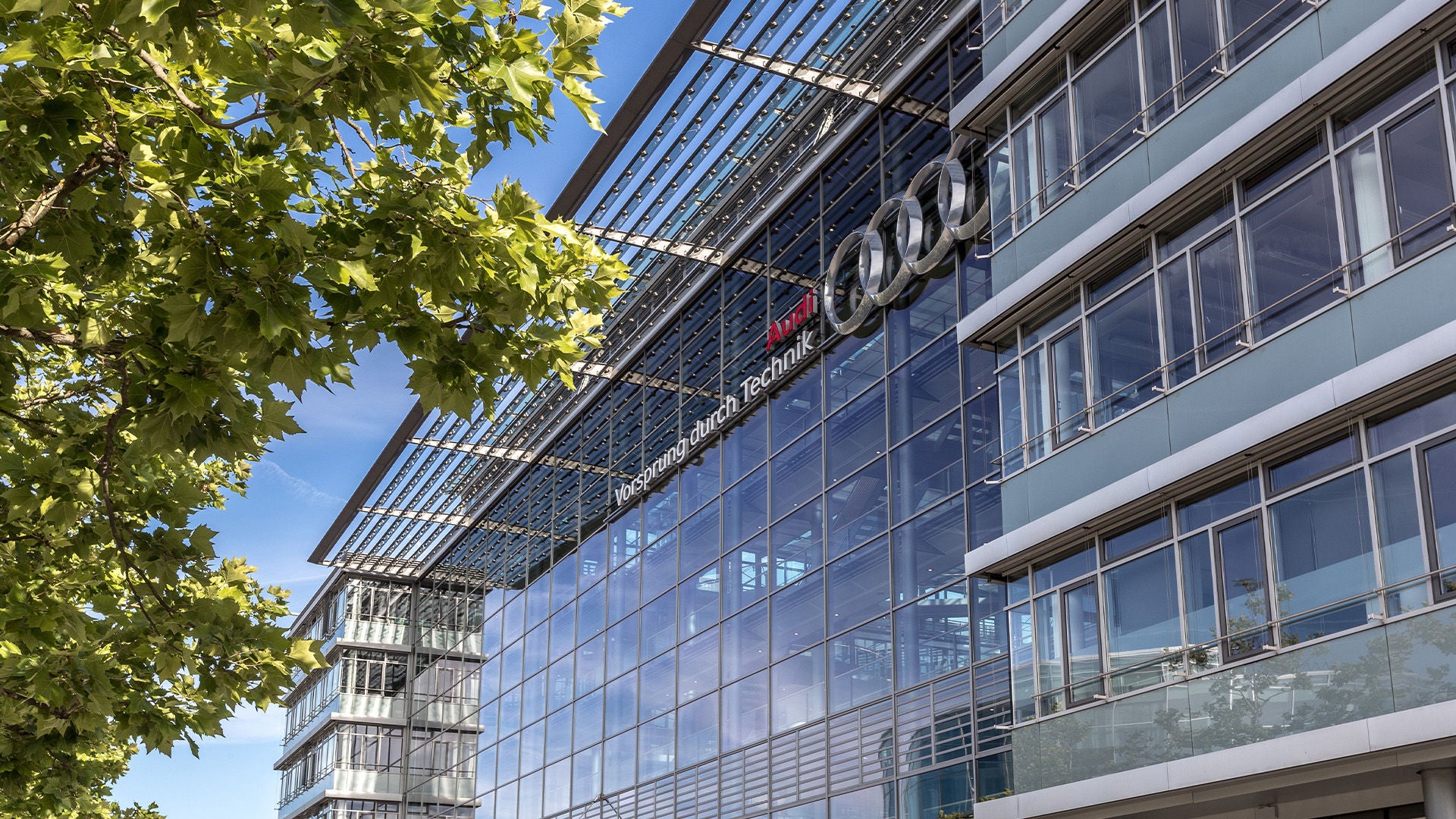 Glass façade of an Audi building with logo and the slogan “Vorsprung durch Technik” in sunny weather.