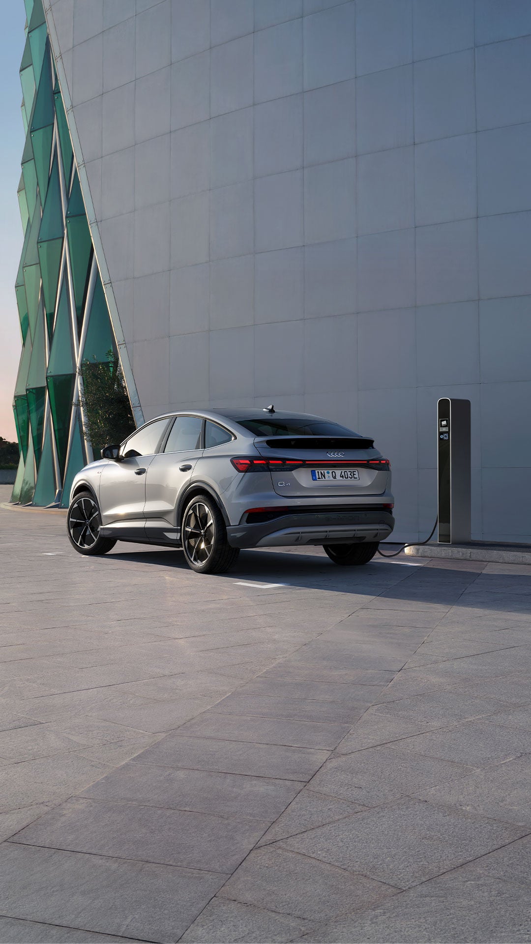 Silberner Audi Q4 Sportback e-tron mit Sportfelgen lädt an einer Ladesäule vor modernem Gebäude bei Sonnenuntergang.
