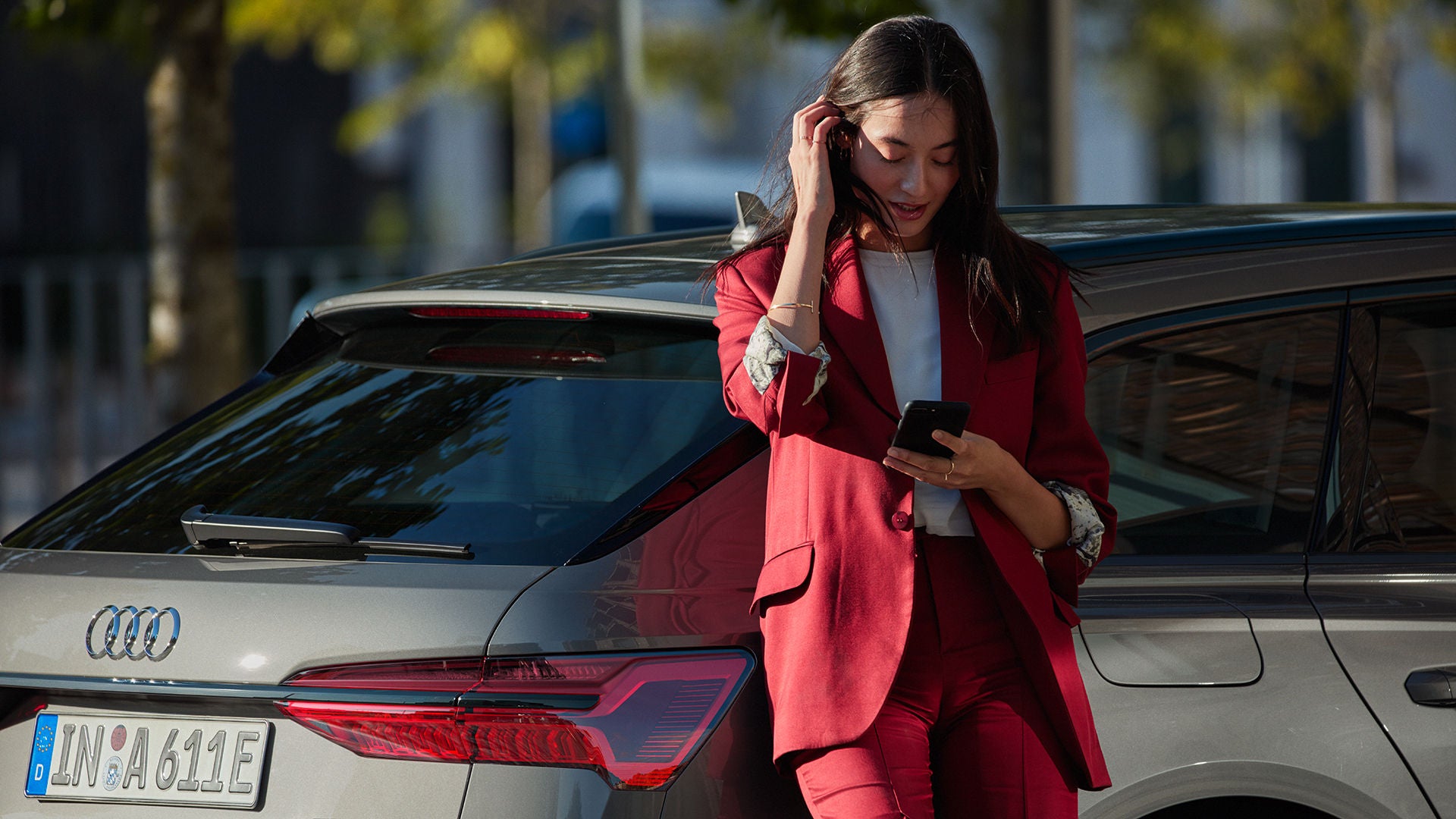 En kvinna med en mobil i handen som står lutad mot bakre delen av en mörkgrå Audi A6 TFSI e 