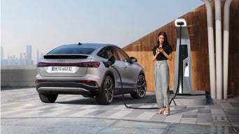 A silver Audi Q4 Sportback e-tron is parked at a charging station, with a woman using her phone nearby against a modern urban backdrop.