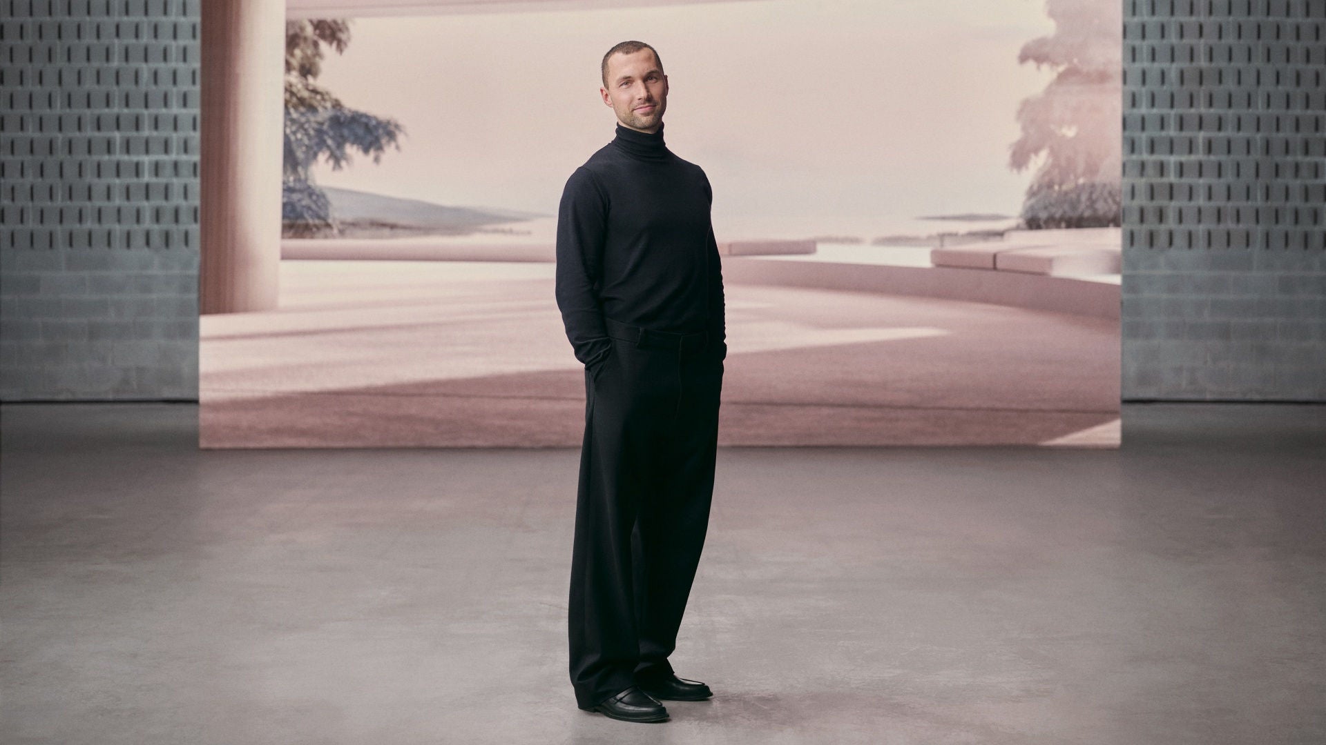 Mann in schwarzem Rollkragenpullover steht lächelnd in modernem Raum mit großem Bildschirm im Hintergrund.