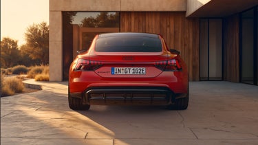 Heckansicht eines roten Audi e-tron GT quattro, der im Licht eines Sonnenuntergangs auf der gepflasterten Auffahrt vor einem Haus mit moderner Architektur steht.