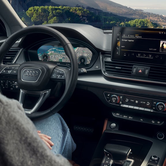 Driver's perspective of an Audi interior with steering wheel, digital displays, and a multimedia system.