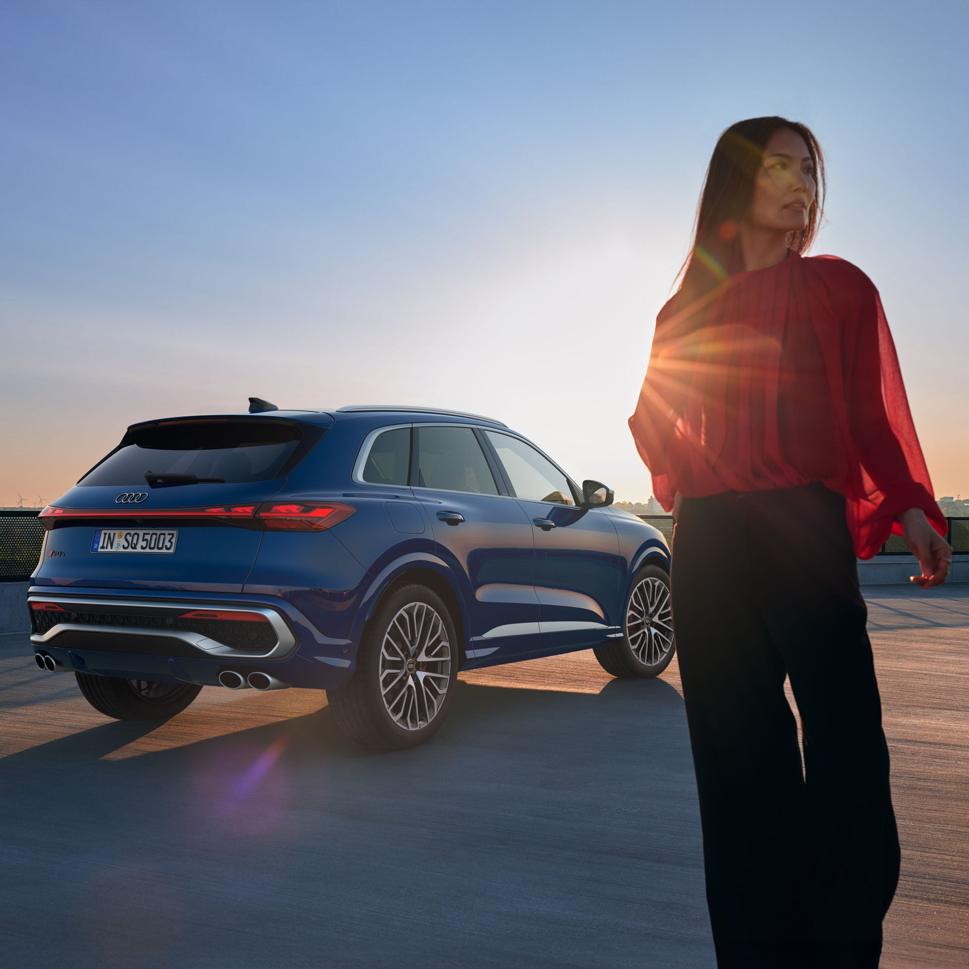 Frau in roter Bluse geht bei Sonnenuntergang an einem blauen Audi SQ5 vorbei; Sonnenlicht fällt durch das Bild.