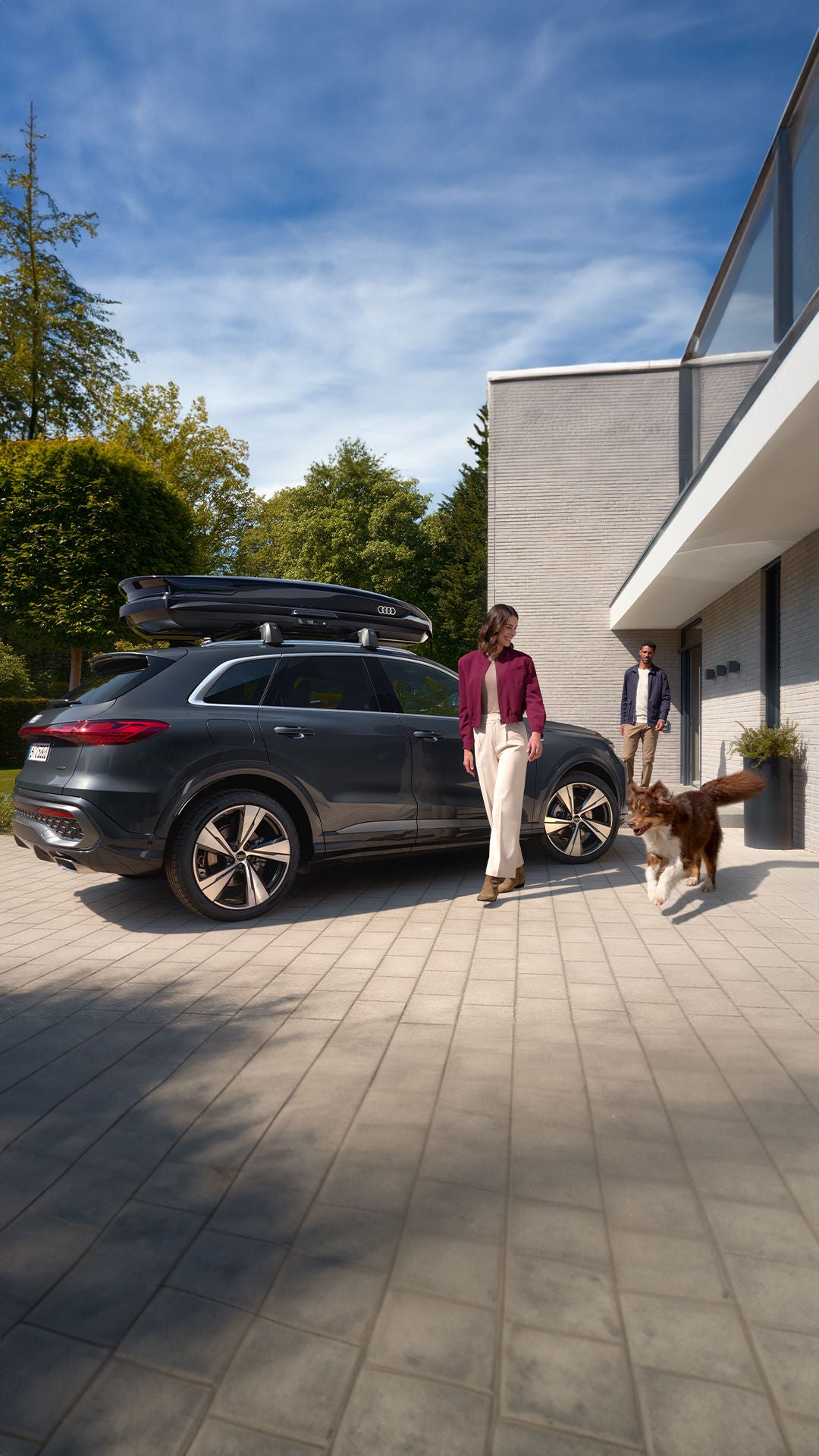 Un Audi Q5 gris avec un coffre de toit installé est garé devant une maison moderne, accompagné d’un couple et de leur chien.