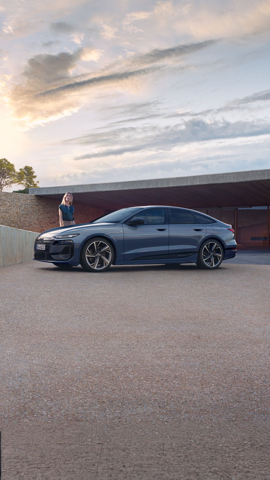 Der A6 Sportback e-tron steht im Seitenprofil vor dem Carport eines Hauses. Daneben steht eine junge Frau.