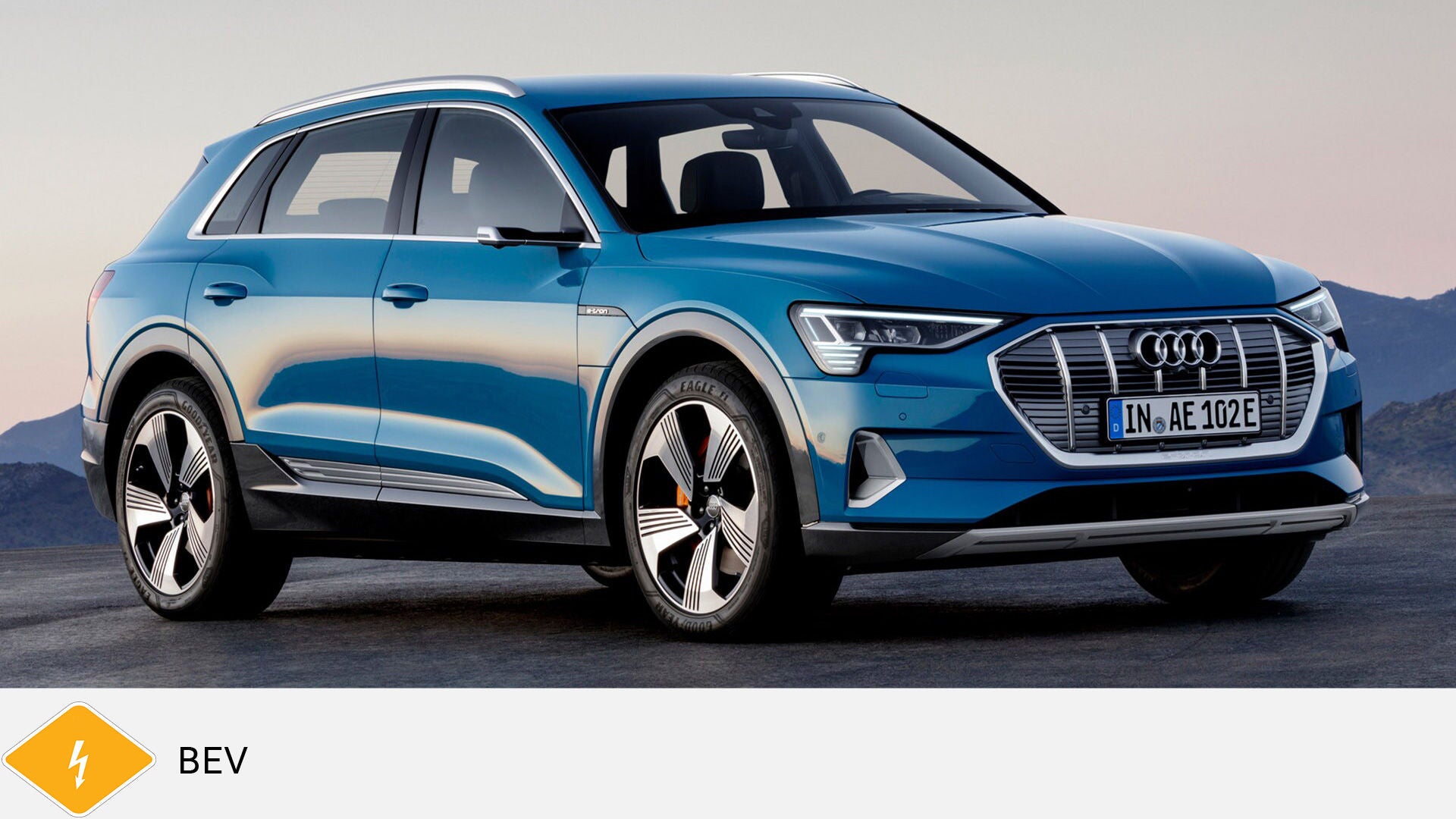 Blue Audi e-tron SUV on asphalt in front of a mountain panorama, diagonally from the front. Yellow BEV symbol with lightning bolt at the bottom.