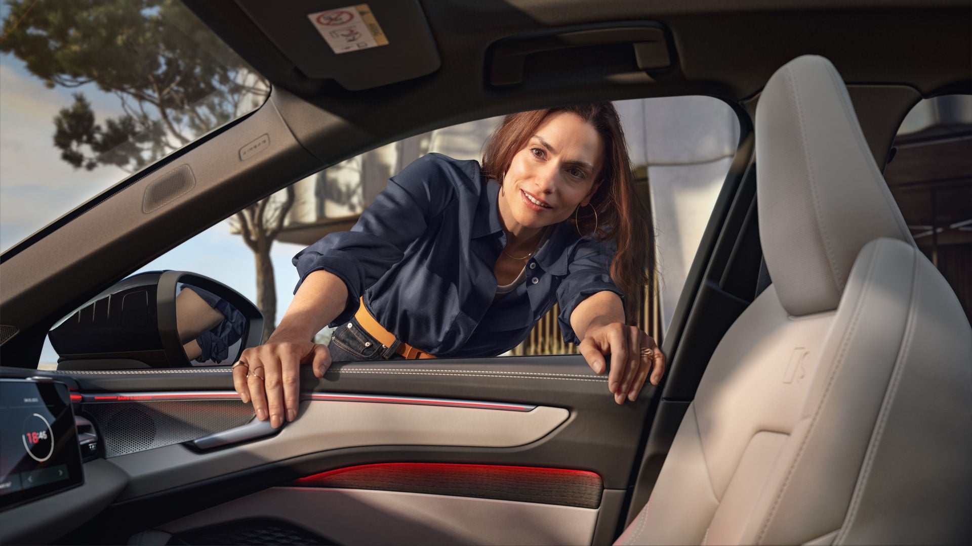 Eine Frau beugt sich durch die geöffnete Tür in den Innenraum eines Autos und hält sich mit beiden Händen am Türrahmen fest.
