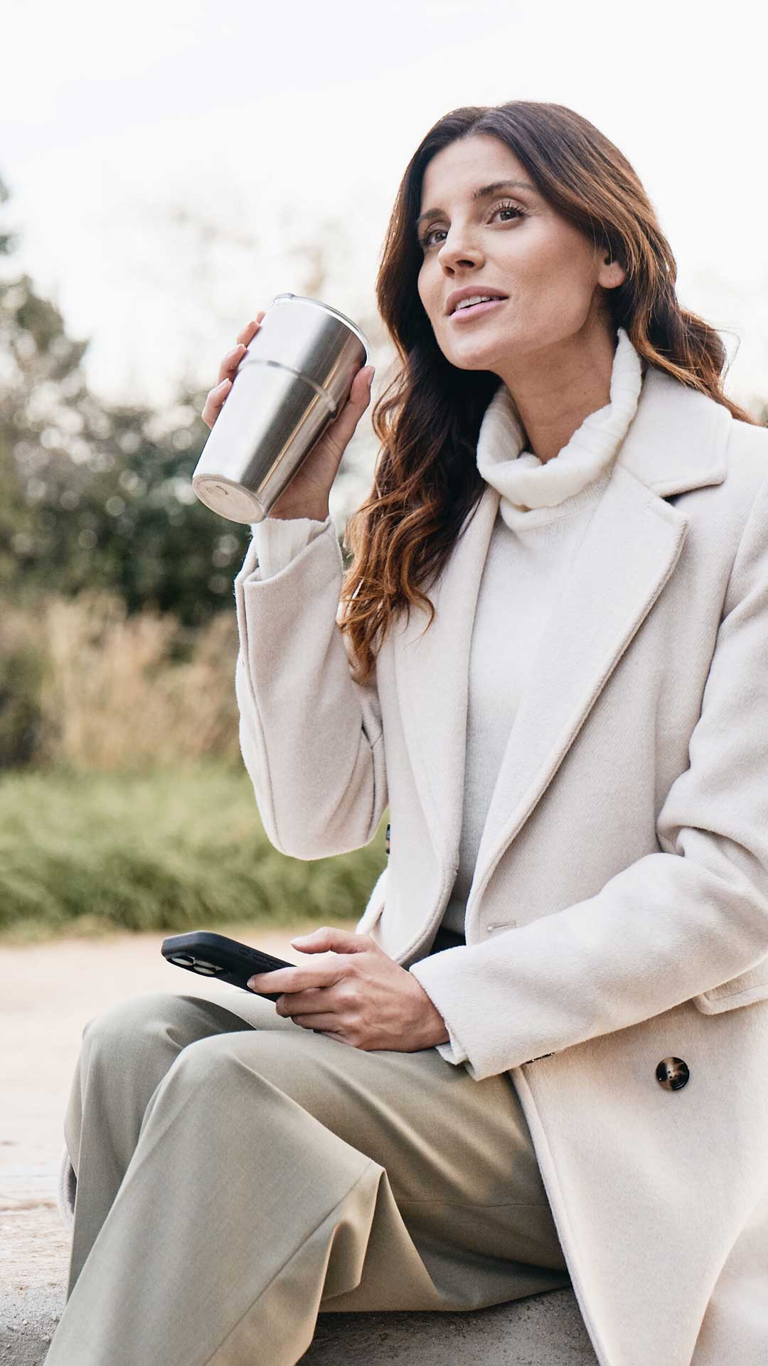 Eine Frau mit weißem Mantel hält in er rechten Hand einen Trinkbecher, in der linken ein Smartphone. Im Hintergrund erkennt man unscharf einen Audi Avant. 