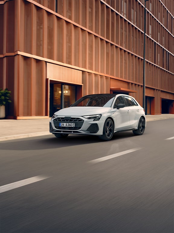 Weißer Audi A3 Sportback TFSI e fährt durch städtische Umgebung mit moderner Architektur.