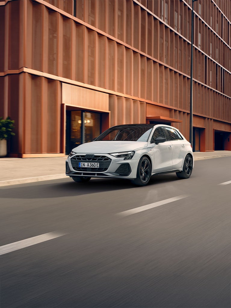 Weißer Audi A3 Sportback TFSI e fährt durch städtische Umgebung mit moderner Architektur.