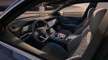 Blick in das Cockpit des SQ8 SUV von der Fahrerseite, im Hintergrund ein futuristisches Gebäude mit geschwungener Form. Vor dem Fahrzeug steht eine modisch gekleidete junge Frau.