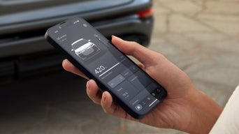 De hand van een vrouw reikt van rechts naar de foto. Ze houdt een smartphone in haar hand, waarvan het display functies toont die de Q6 e-tron biedt. Een groene Audi Q6 e-tron staat wazig op de achtergrond.