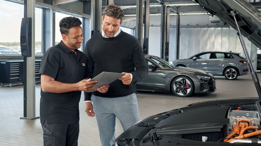 Audi-Mitarbeiter und Kunde betrachten gemeinsam ein Tablet in einer Werkstatt neben einem geöffneten Fahrzeugmotor.