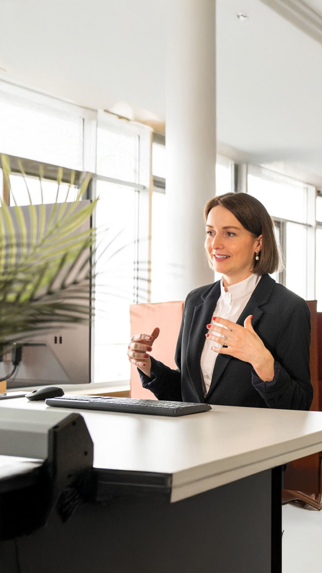 Employee is standing at her desk and colleagues are chatting in the background