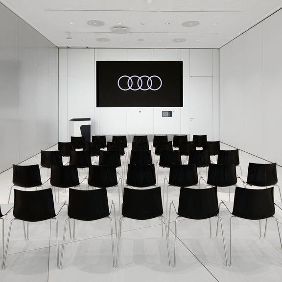 Kleiner, weißer Vortragssaal mit schwarzer Bestuhlung und Audi Logo auf großem Bildschirm.