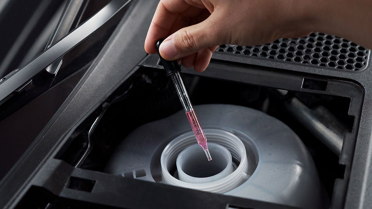 Hand hält Pipette mit rosa Flüssigkeit über geöffnetem Einfüllstutzen im Motorraum eines Fahrzeugs zur Flüssigkeitskontrolle.