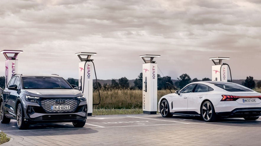 Zwei Audi Elektrofahrzeuge laden an Ionity-Ladesäulen auf einem Parkplatz bei bewölktem Himmel.
