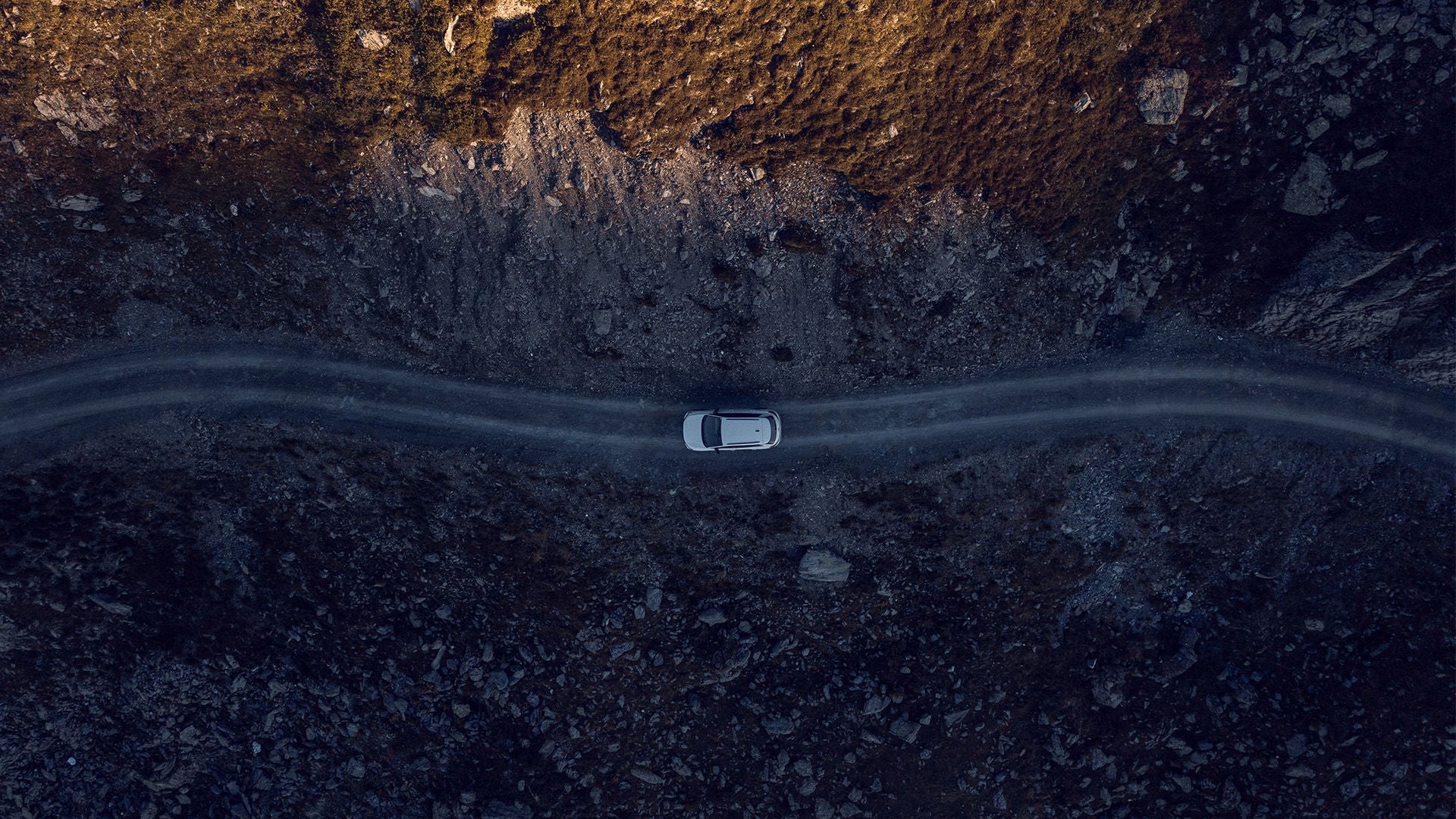 Luftaufnahme eines Autos auf schmaler Straße in felsiger Landschaft bei tiefstehendem Sonnenlicht.          