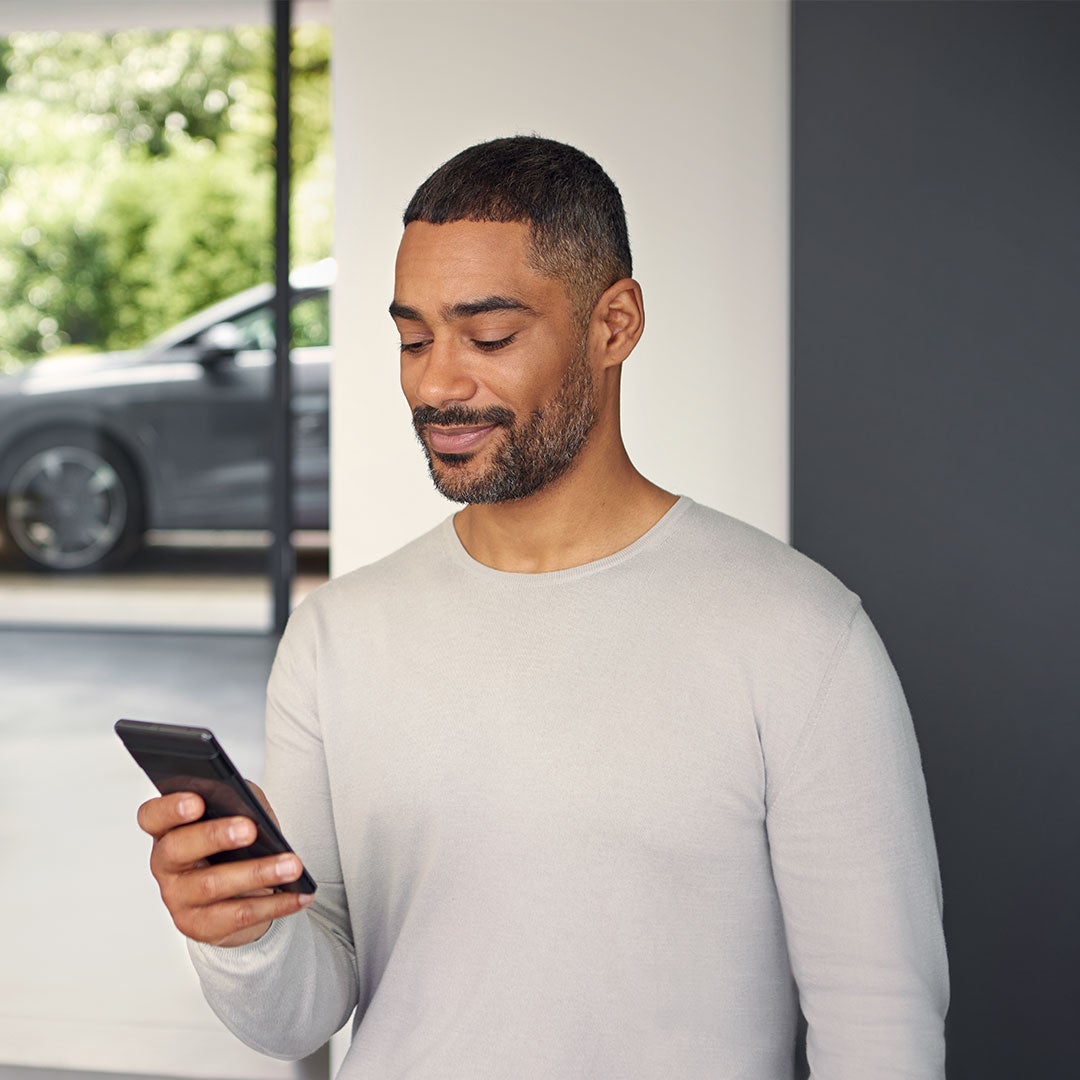 Un homme en pull beige tient un téléphone portable à la main et regarde quelque chose sur celui-ci. En arrière-plan, une voiture est garée à l'extérieur de la maison dans laquelle se trouve l'homme.