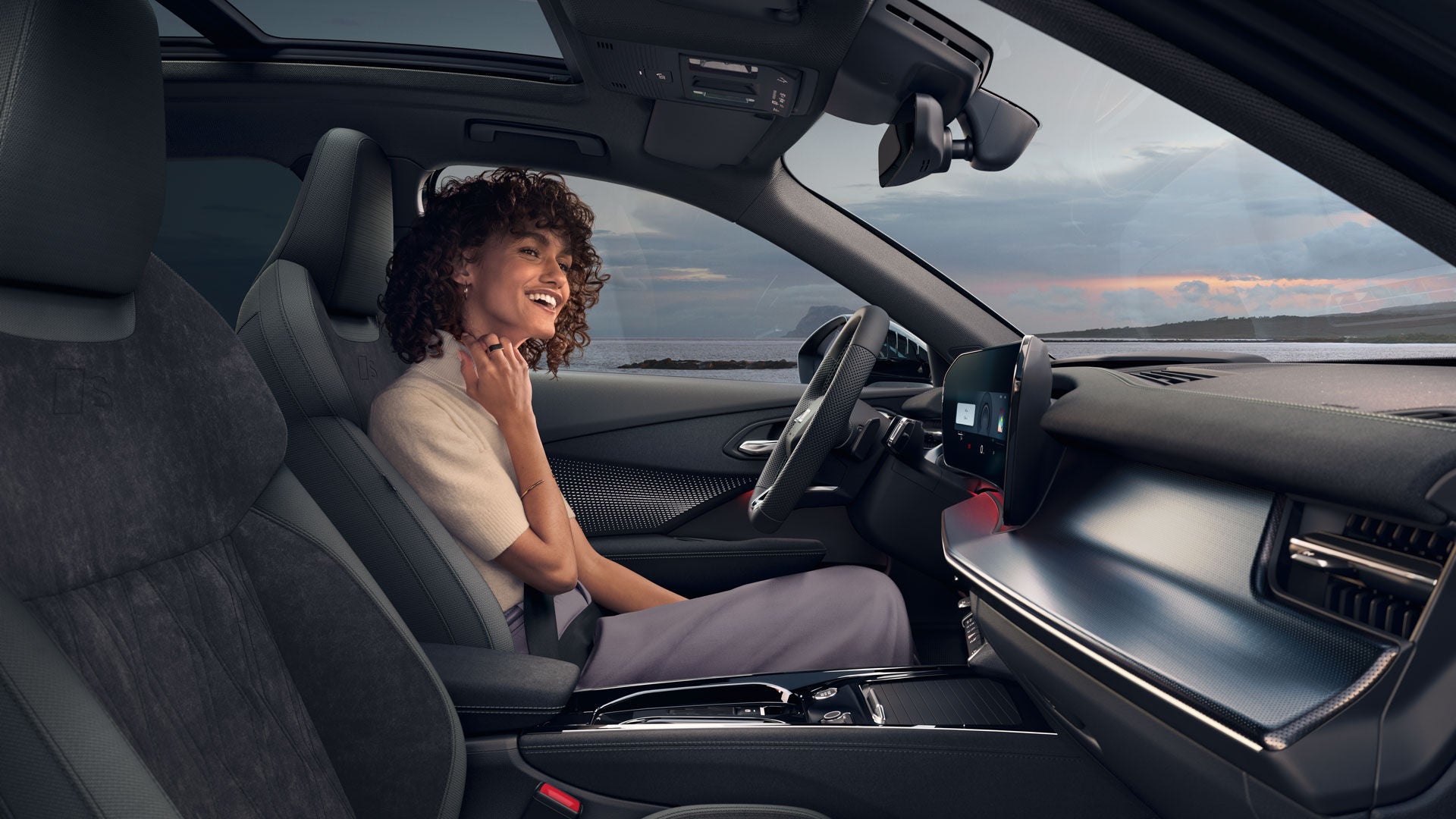 Eine Frau mit lockigen Haaren sitzt lachend am Steuer eines Audi. Durch die Frontscheibe ist ein Sonnenuntergang zu sehen.