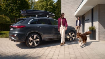 Dunkler Audi-SUV mit Dachbox vor modernem Haus; Frau läuft mit Hund, Mann steht an der Haustür.