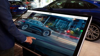 Person bedient einen großen Touchscreen mit Audi-Fahrzeugdarstellung, im Hintergrund mehrere Autos im Ausstellungsraum.