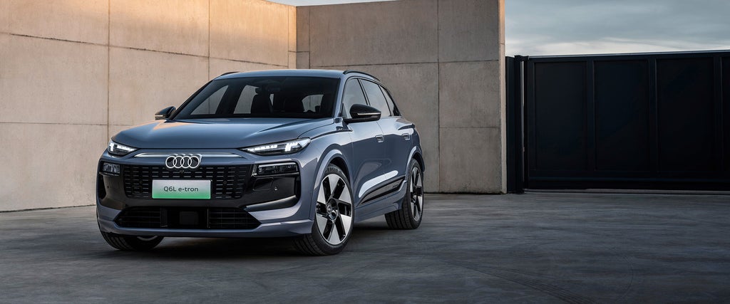 Ein Audi Q6L e-tron in metallic-blauer Lackierung steht vor einer modernen Betonwand und einem schwarzen Tor, während der Himmel in sanften Abendfarben leuchtet.