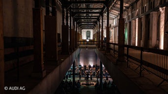 Orchester mit Dirigent spielt in einer großen Industriehalle; Publikum sitzt davor, dramatische Beleuchtung, dunkle Stahlträger.