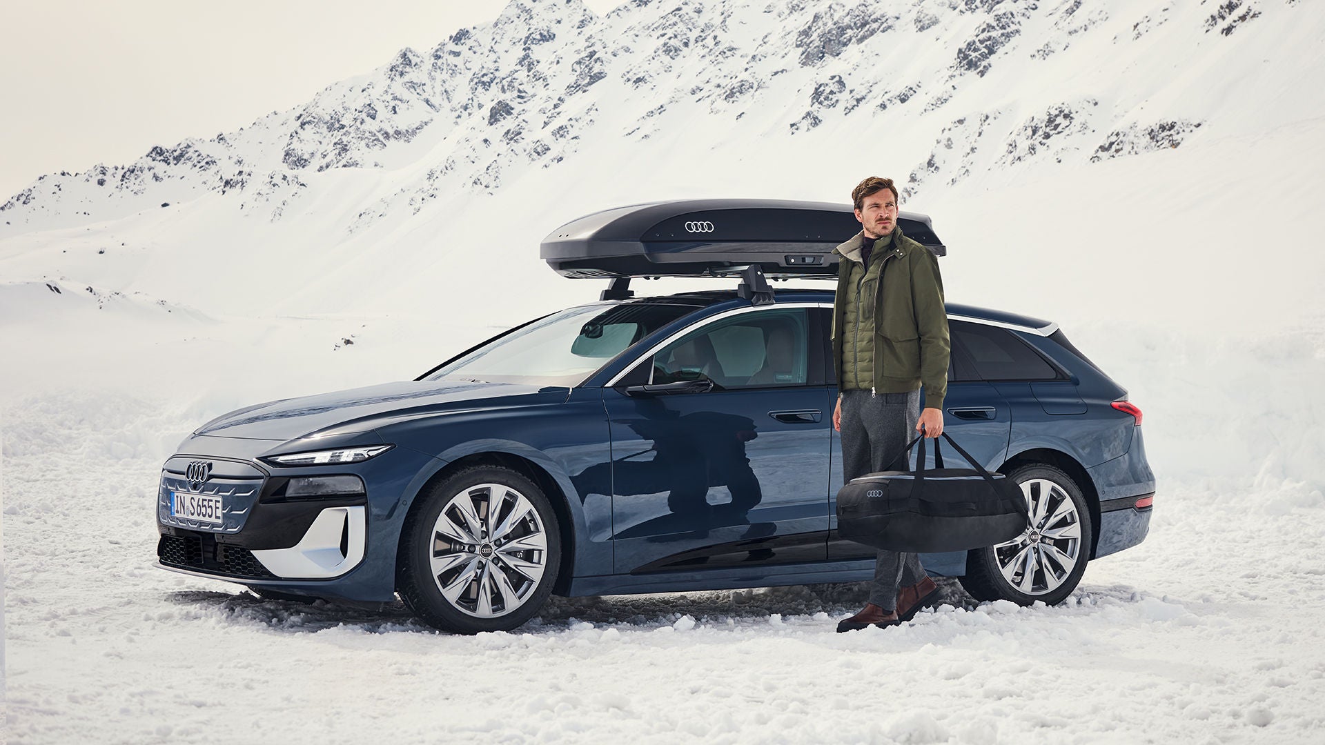 Ένας άντρας στέκεται σε ένα χιονισμένο τοπίο μπροστά από ένα Audi και κρατάει μια τσάντα του σκι.