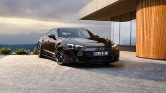 A black Audi S e-tron GT parked on a modern stone driveway next to a contemporary wooden building and scenic landscape.