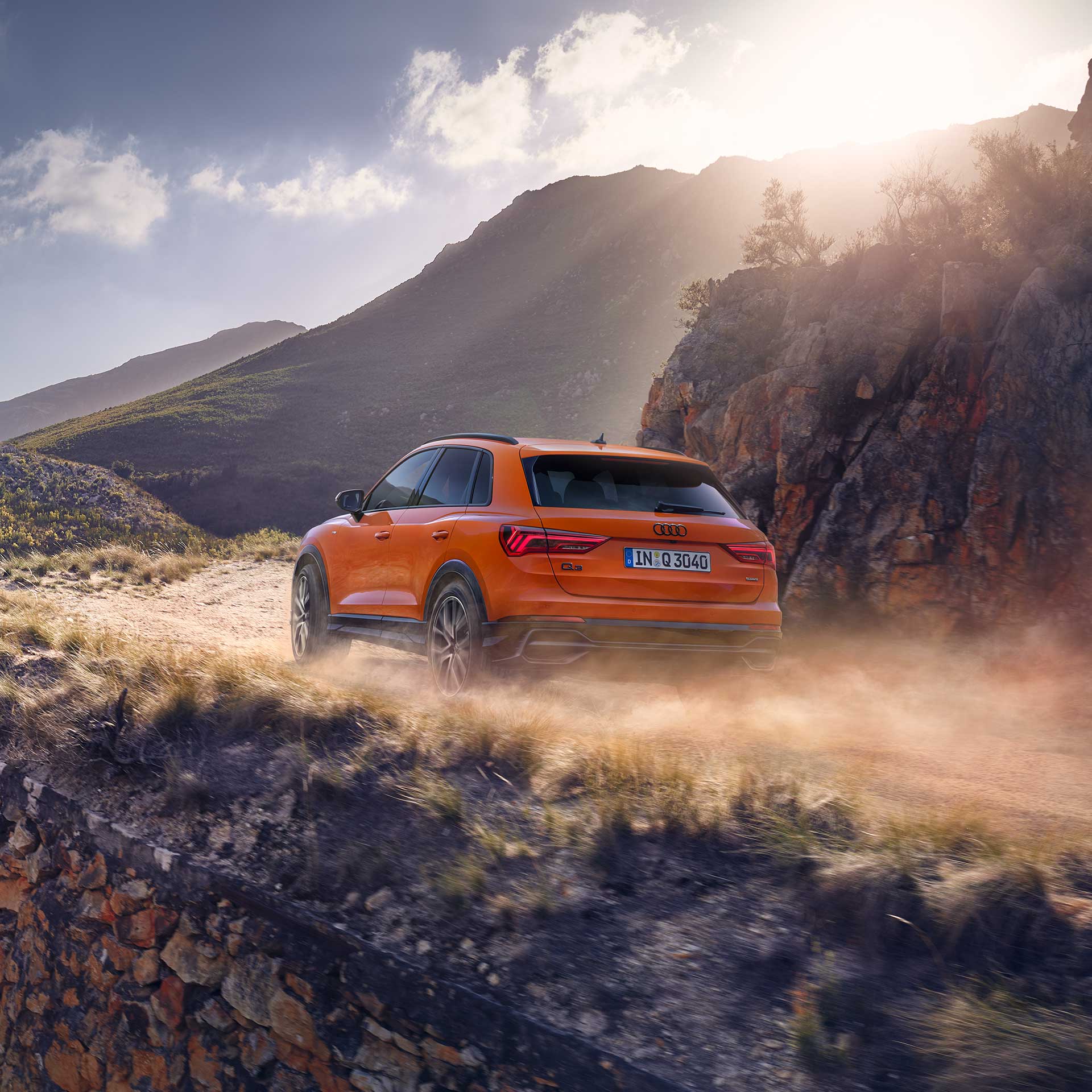 Ein orangener Audi fährt auf einer hügeligen, staubigen Landstraße und im Hintergrund sind Berge und die Sonne.