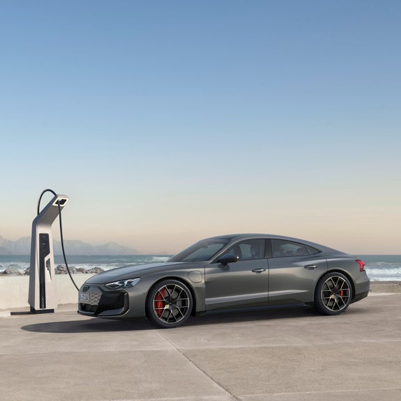 Ein grauer Audi RS e-tron GT lädt an einer Ladestation an der Küste mit Wellen und Bergen im Hintergrund.