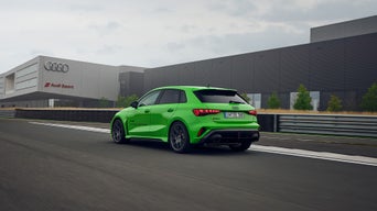 Light green Audi RS 3 Sportback on a training ground, in the background is a building with the Audi Sport logo.
