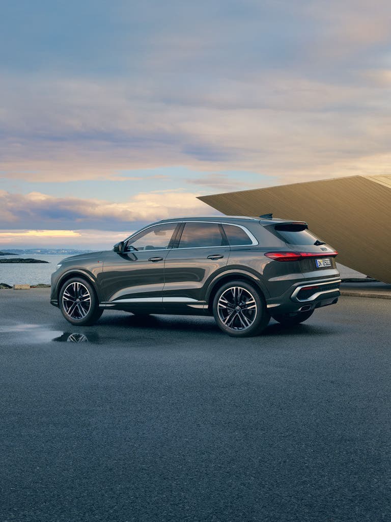 Ein grauer Audi Q5 SUV steht auf einer Parkfläche mit Meerblick und moderner Architektur im Hintergrund.