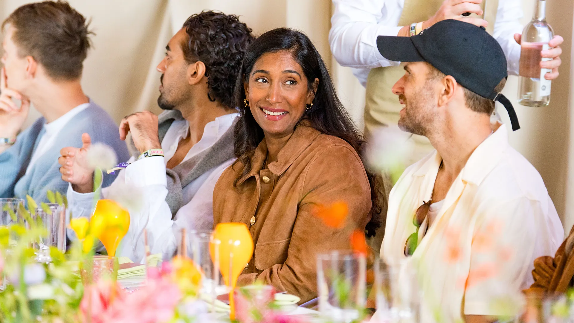A woman in a tan suede jacket smiles over her shoulder at a dinner table decorated with bright orange and yellow flowers, surrounded by other guests. A woman in a tan suede jacket smiles over her shoulder at a dinner table decorated with bright orange and yellow flowers, surrounded by other guests.