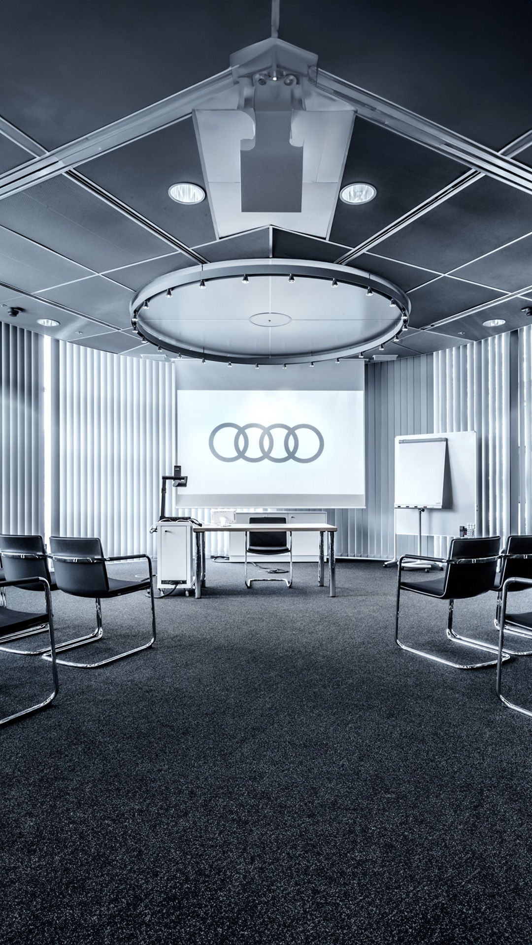 Heller Konferenzraum mit U-förmigem Tisch, schwarzen Stühlen und Audi-Logo auf einem Bildschirm.