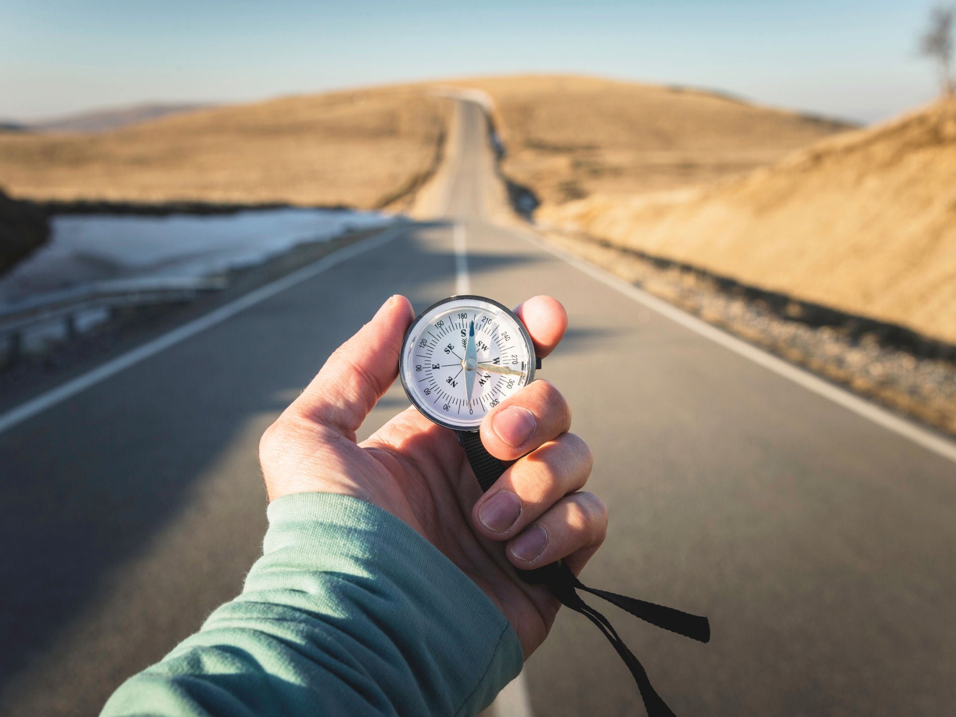One hand holds a compass pointing south. In the background, a long road can be seen in a desert landscape.