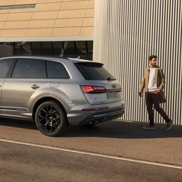 A person walking past a parked silver Audi Q7 tfsie on a sunny day.