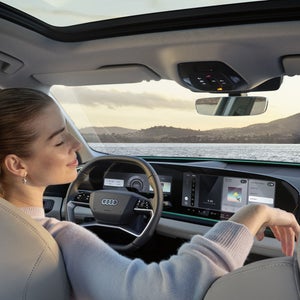 Interiören i Q6 SUV e-tron sett från baksätet. En ung kvinna sitter i förarsätet och lutar sig tillbaka med slutna ögon. Genom vindrutan kan man se havet och bergen.