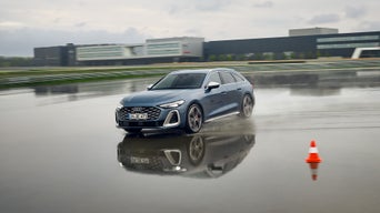 Audi S5 Avant driving through a deep puddle on a wet training ground, with an Audi building in the background.