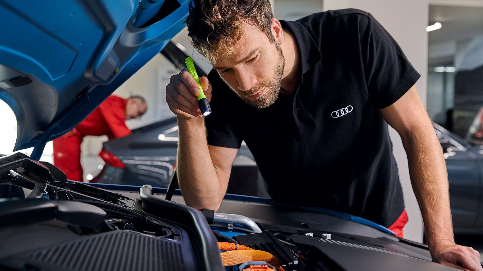 Audi-Mitarbeiter inspiziert mit Taschenlampe den Motorraum eines Fahrzeugs in einer Werkstattumgebung.