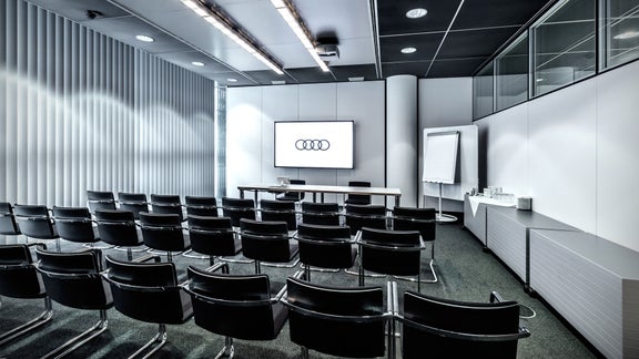 Kleiner Präsentationsraum mit Reihen schwarzer Stühle, Audi-Logo auf einem Bildschirm und einem Buffettisch.