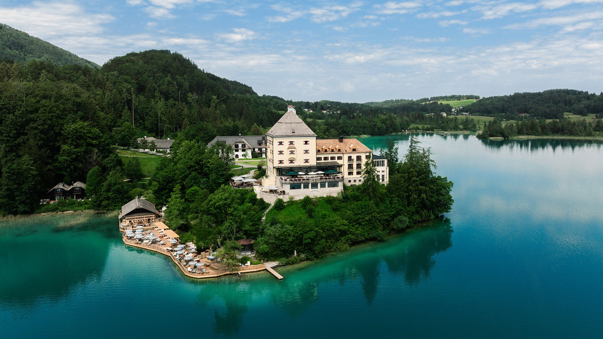 Luftaufnahme eines Schlosses auf einer Halbinsel in türkisblauem See, umgeben von Wald und Hügeln.