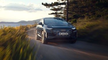 De Audi Q4 SUV e-tron, van voren in een schuine hoek gezien, terwijl hij over een landweg langs een bosmeer raast, met op de achtergrond een bergachtig landschap.