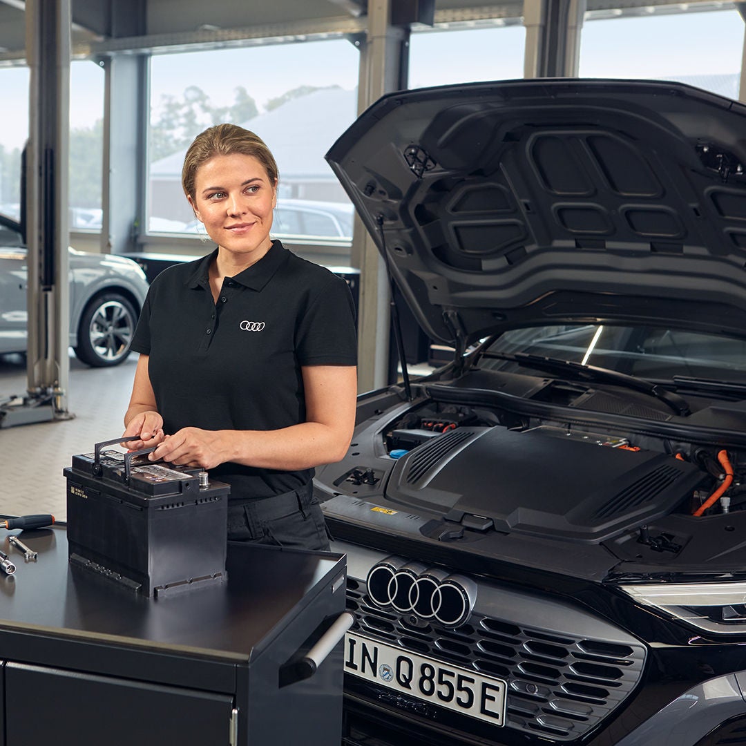 Audi-Mitarbeiterin steht vor geöffneter Motorhaube und hält eine Starterbatterie in der Werkstatt.