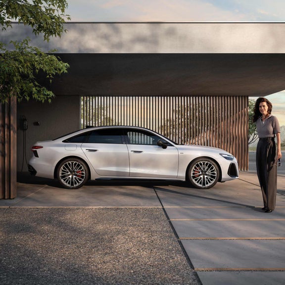 Die A6 Limousine e-hybrid steht seitlich vor einer Hauswand, an welche eine Ladestation montiert ist. Vor dem Auto steht eine junge Frau.