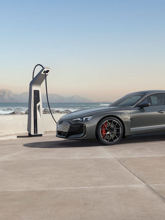 Ein grauer Audi RS e-tron GT lädt an einer Ladestation an der Küste mit Wellen und Bergen im Hintergrund.